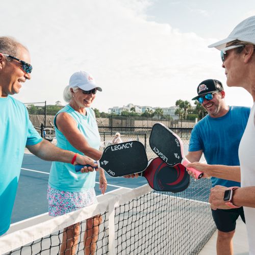 Four people on a pickleball court are tapping paddles together over the net, smiling.