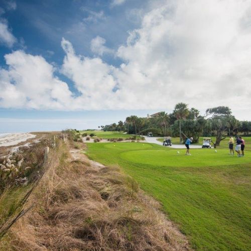 A coastal golf course with a group of golfers on the green, golf carts nearby, and ocean waves lapping at the shore under a partly cloudy sky.