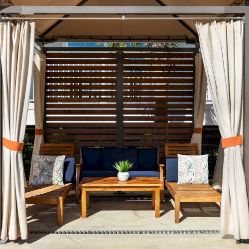 A cozy outdoor seating area with wooden furniture, cushions, curtains, and a potted plant on a table under a canopy.