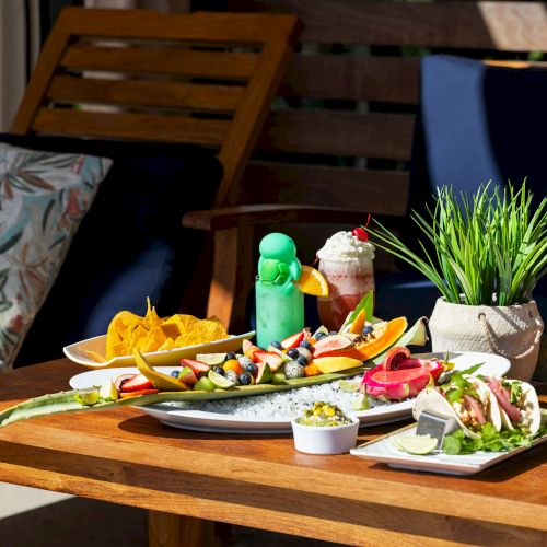 A wooden table with assorted fruits, chips, drinks, tacos, and a potted plant. Cozy seating with a floral cushion is shown in the background.