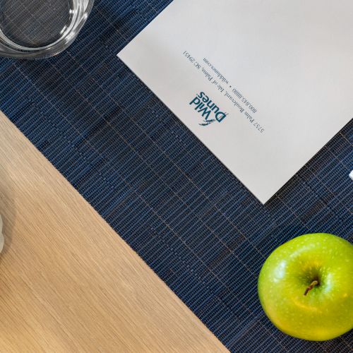 A green apple, glass of water, plant, pen, and paper with "Waldhotel" printed, are arranged on a blue placemat and wood table.