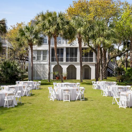 An outdoor event setup with round tables and chairs on a grassy area, surrounded by palm trees and buildings in the background.