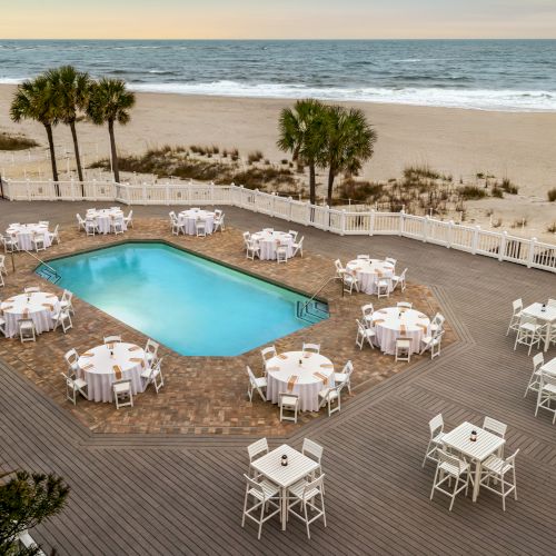 A beachside area with a pool is surrounded by tables and chairs on a wooden deck near the ocean under a serene sky.