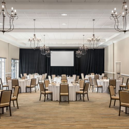 The image shows a banquet hall with round tables and chairs, white tablecloths, and chandeliers, set up for an event with a projector screen.