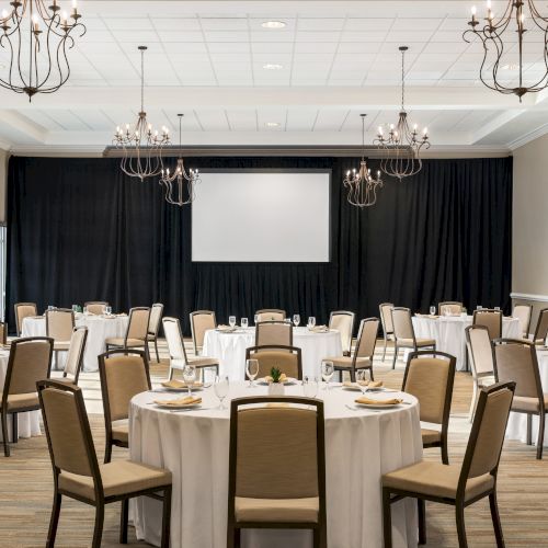 A banquet hall with round tables covered in white tablecloths, chairs, chandeliers, and a large screen at the front, set for an event.