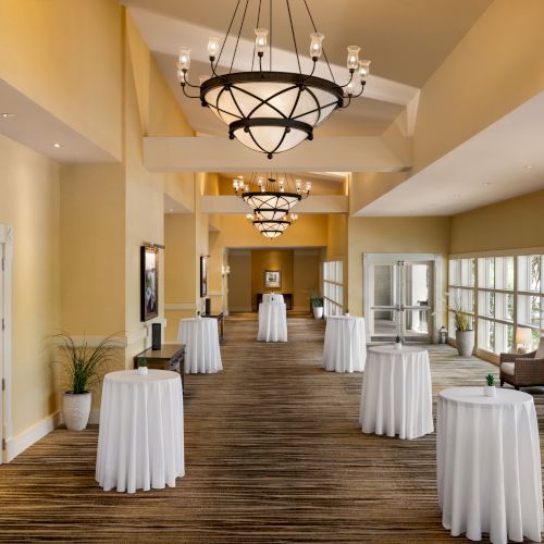 A spacious hallway with chandeliers, round tables with white cloths, and seating areas by large windows, exuding an elegant ambiance.