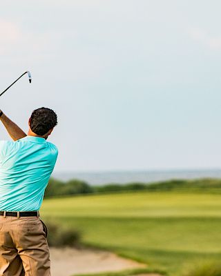 A person is playing golf, taking a swing on a green course near the sea with a flag in the background.