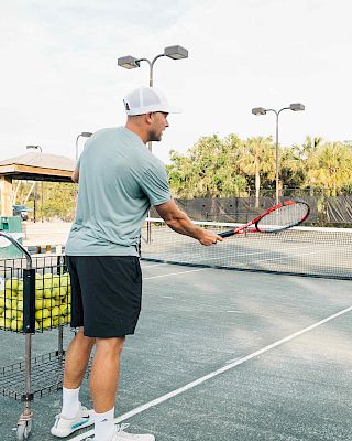 A man on a tennis court is practicing with a basket full of tennis balls, preparing to hit a shot.