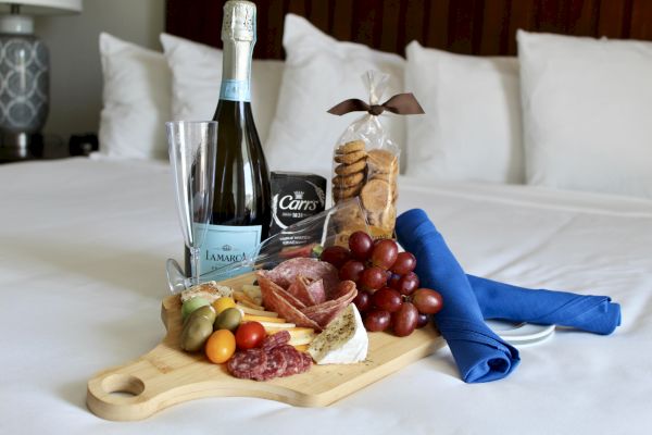 A cheese board with salami, grapes, olives, and tomatoes, accompanied by champagne, cookies, and a blue napkin on a bed.