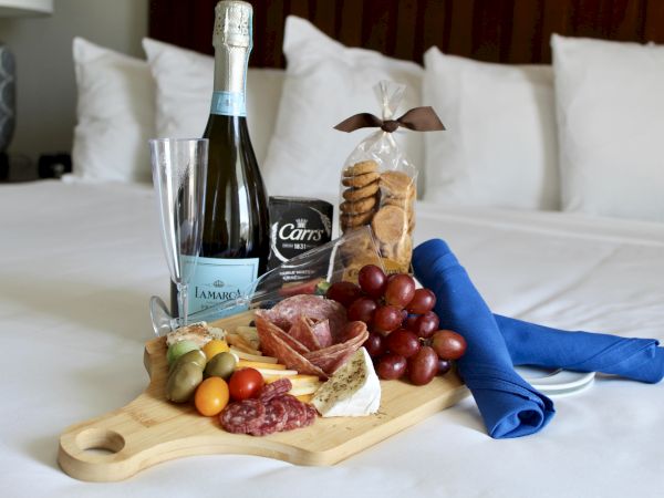 A cheese board with salami, grapes, olives, and tomatoes, accompanied by champagne, cookies, and a blue napkin on a bed.