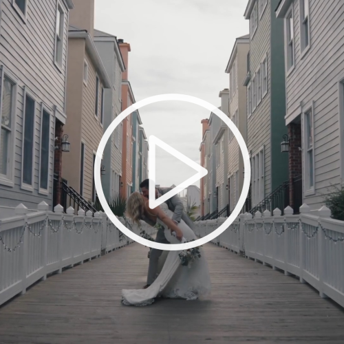 A couple is embracing on a walkway lined with festive lights, between colorful houses, overlaid with a play button.
