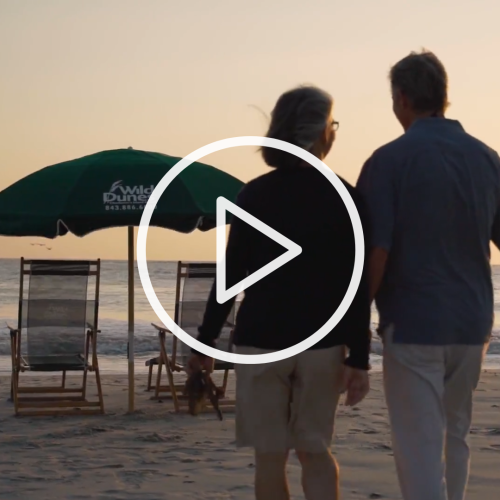 Two people walk on a beach at sunset, heading towards chairs under a green umbrella.