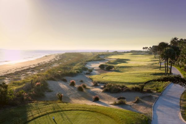 A scenic coastal golf course with well-maintained greens, adjacent to a beach, under a clear sky with a sun setting or rising.