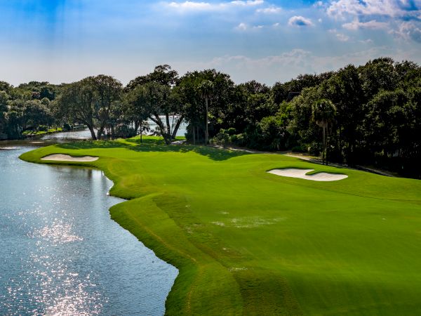 A lush golf course fairway beside a calm waterway, with sand bunkers and mature trees lining the scenic green landscape.