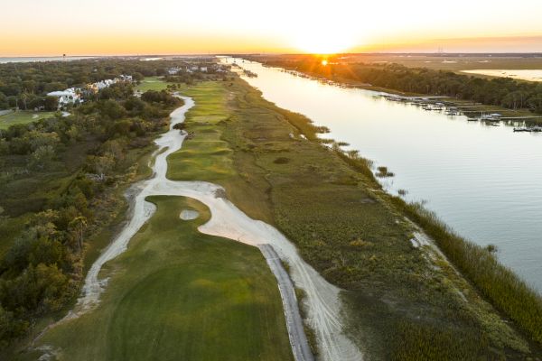 Aerial view of a golf course next to a river at sunset, with a winding path, greens, and trees surrounding the area.