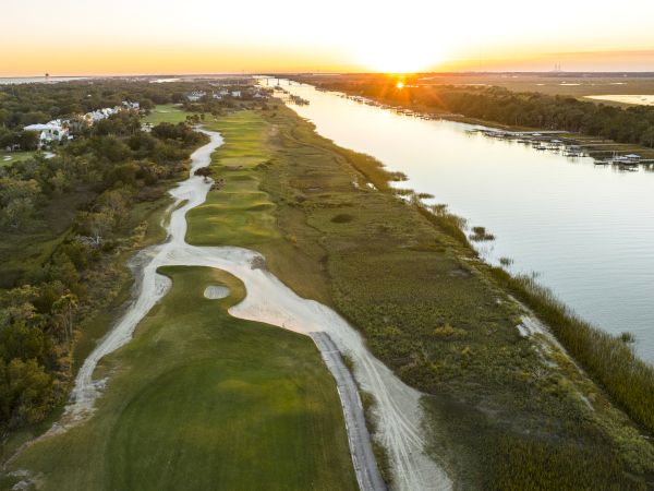 Aerial view of a golf course next to a river at sunset, with a winding path, greens, and trees surrounding the area.