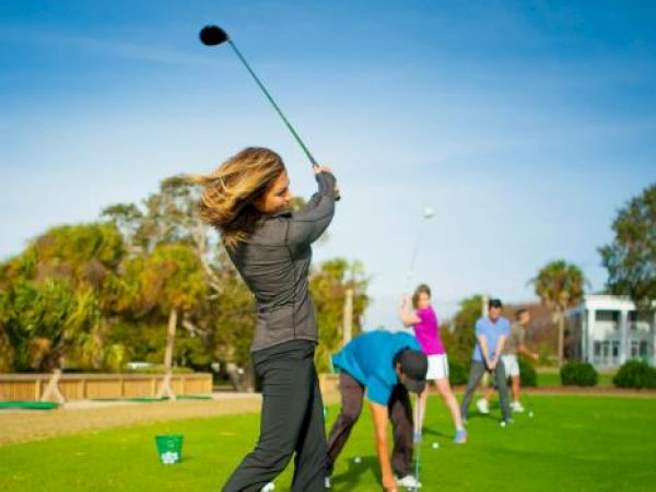People are practicing their golf swings on a sunny day at a driving range with greenery and buildings in the background.