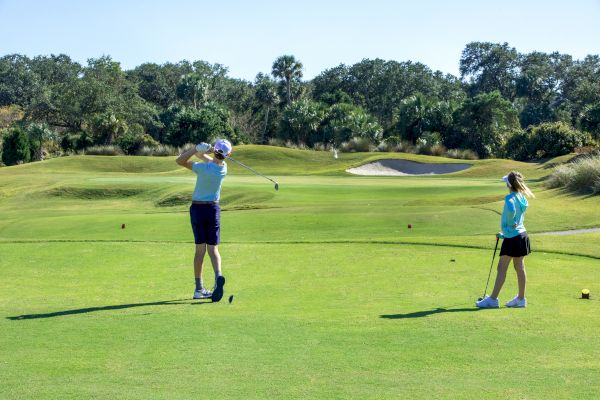 Two people on a golf course, one is swinging a club, and the other is watching. The landscape is green with trees in the background.
