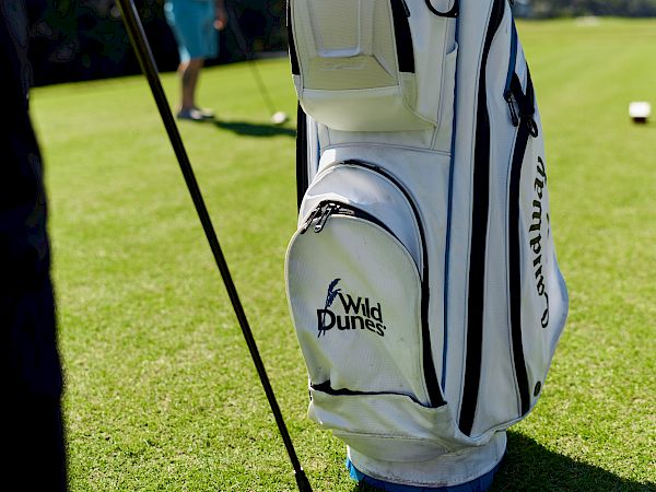 A white golf bag with clubs stands on a sunny golf course, with a person blurred in the background, and a golf ball on the grass.