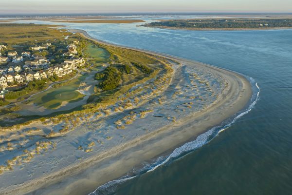 Aerial view of a coastal landscape with houses, golf course, sandy beach, and waves meeting the shore under a vast sky.