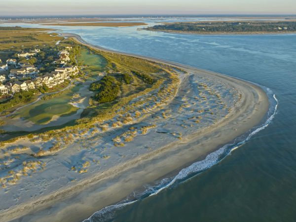 Aerial view of a coastal landscape with houses, golf course, sandy beach, and waves meeting the shore under a vast sky.