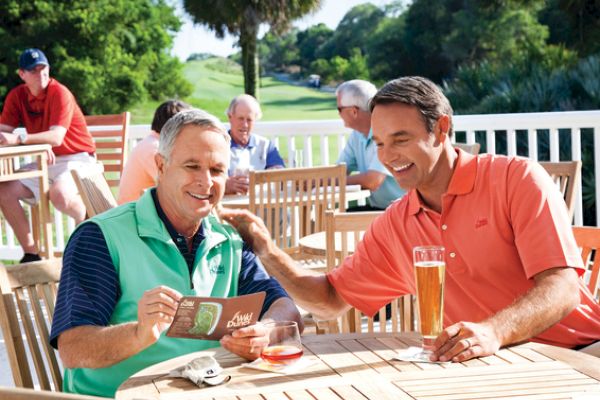 Two men sit at a wooden table outdoors, enjoying drinks and laughing, surrounded by others and greenery.