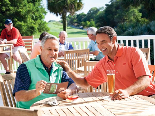Two men sit at a wooden table outdoors, enjoying drinks and laughing, surrounded by others and greenery.