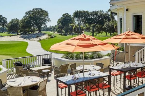 A patio with tables and orange umbrellas overlooks a lush golf course under a clear sky.