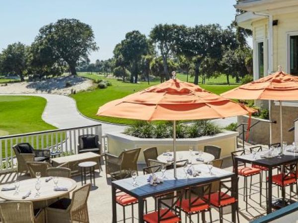 A patio with tables and orange umbrellas overlooks a lush golf course under a clear sky.