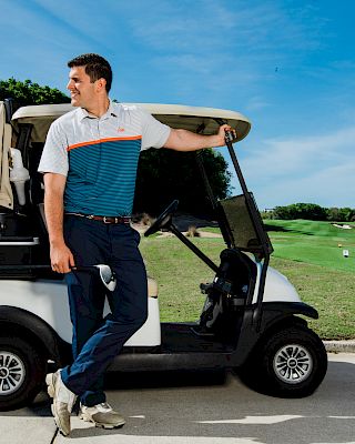 A person wearing a golf outfit leans on a golf cart, looking towards the course, with a grassy field and trees in the background.