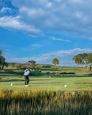 Two people playing golf on a lush course with palm trees under a partly cloudy blue sky.