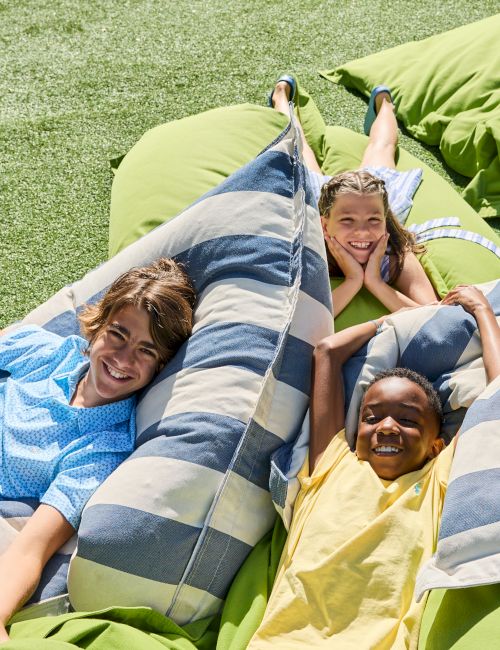 Three kids are relaxing on large striped and green bean bags on grass, smiling and enjoying the sunny outdoors.