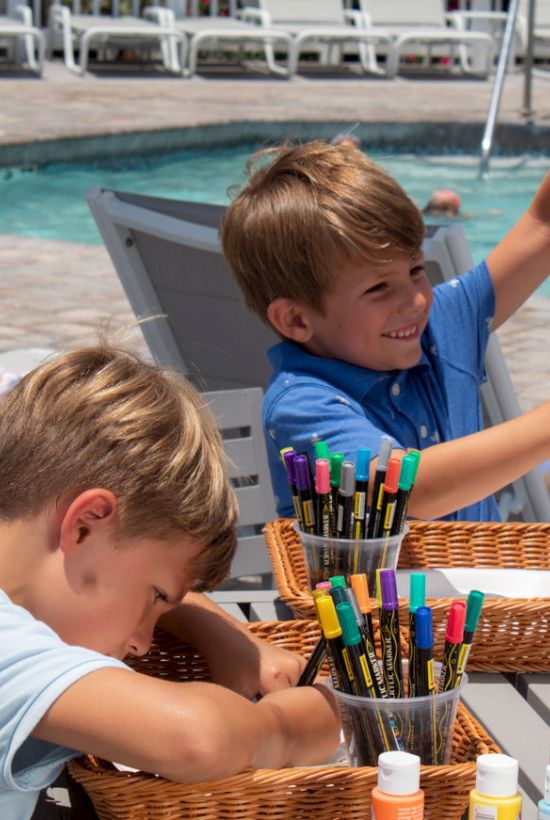 Two children are doing arts and crafts with markers by a pool, enjoying their time outdoors.