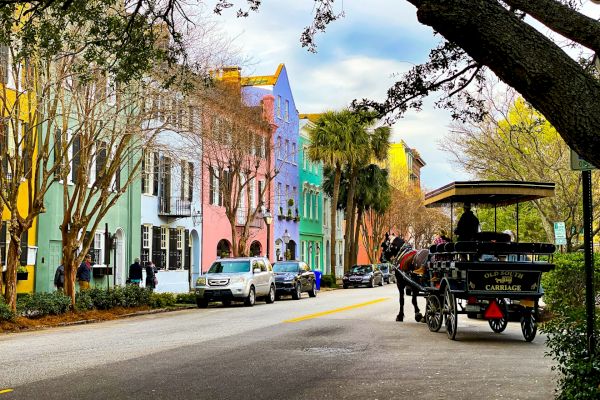Colorful row of historic buildings along a street with a horse-drawn carriage and cars parked, under a canopy of trees.
