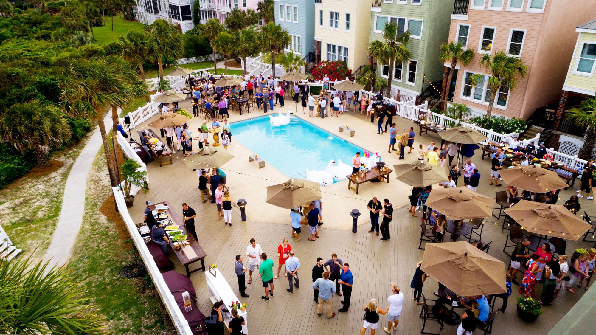 Aerial view of a vibrant outdoor gathering by a pool, with people socializing under umbrellas and colorful buildings in the background.