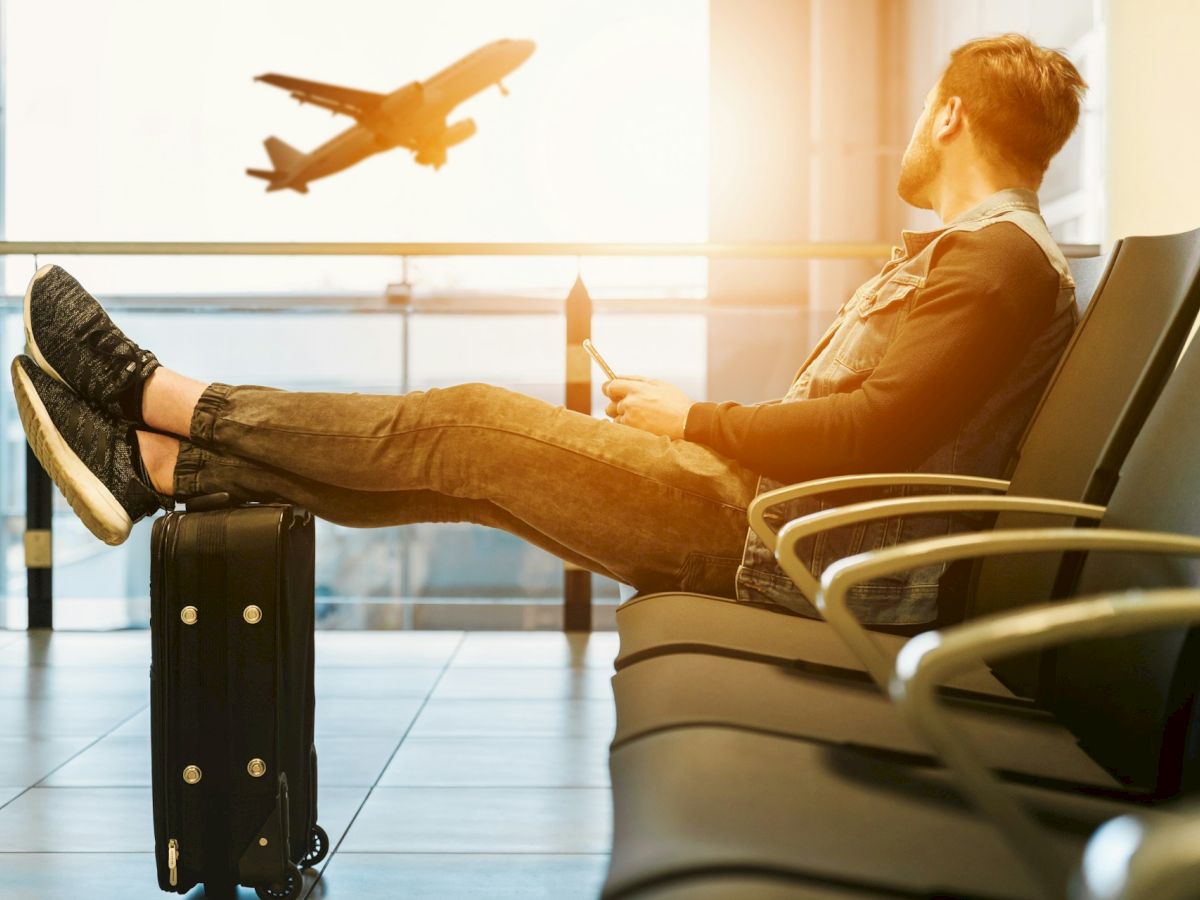 A person sits in an airport lounge, feet on a suitcase, watching a plane take off through large windows.