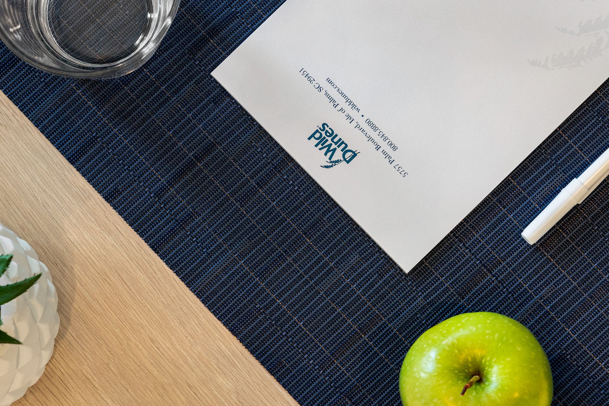 A desk with a water glass, green apple, plant, pen, and a document on a blue woven mat.