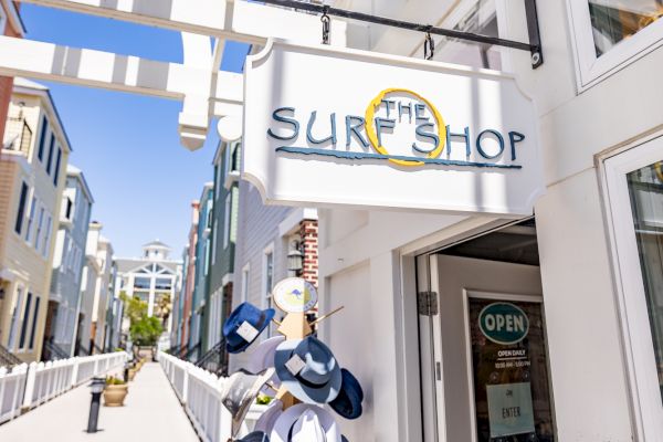 A surf shop entrance with hats displayed outside, located in a bright, sunny area with a trellis structure and boardwalk walkway.