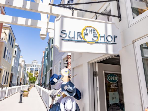 A surf shop entrance with hats displayed outside, located in a bright, sunny area with a trellis structure and boardwalk walkway.