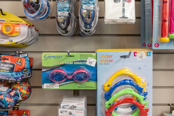 The image shows a retail display with swimming goggles, toy rings, snorkels, and pool accessories on hooks and shelves.