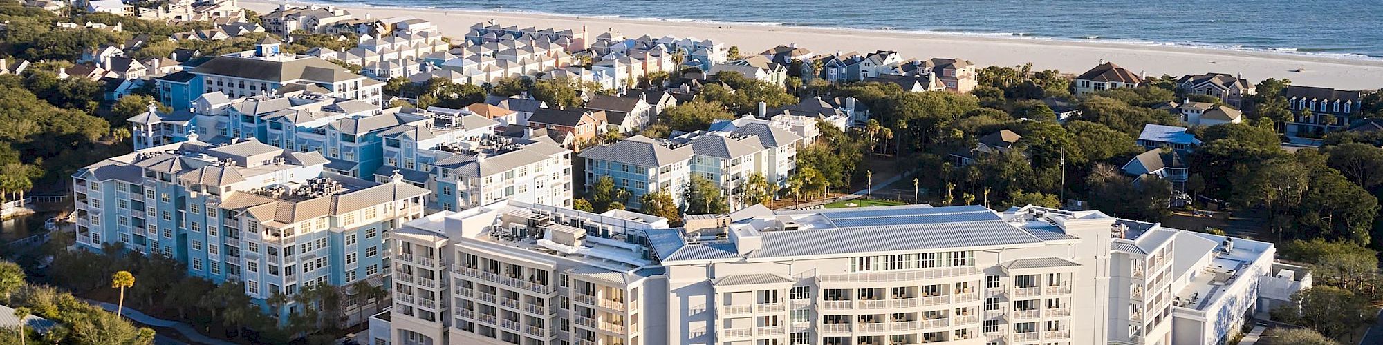 An aerial view of a coastal resort complex with buildings surrounded by trees, overlooking a sandy beach and ocean.