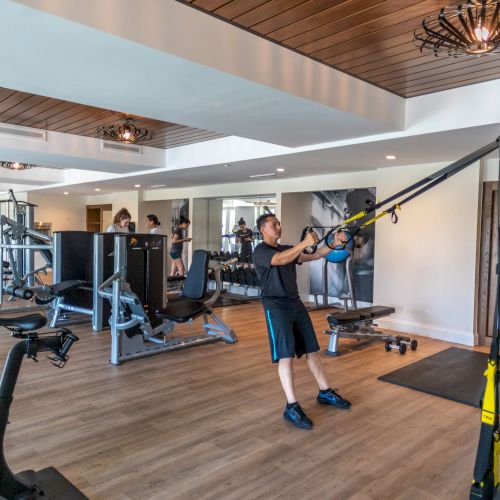 A gym with people exercising using various equipment, including TRX bands and exercise bikes, in a well-lit modern space.