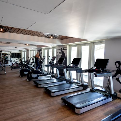 The image shows a gym with treadmills, exercise bikes, and mirrors, featuring wooden flooring and wall graphics of runners.