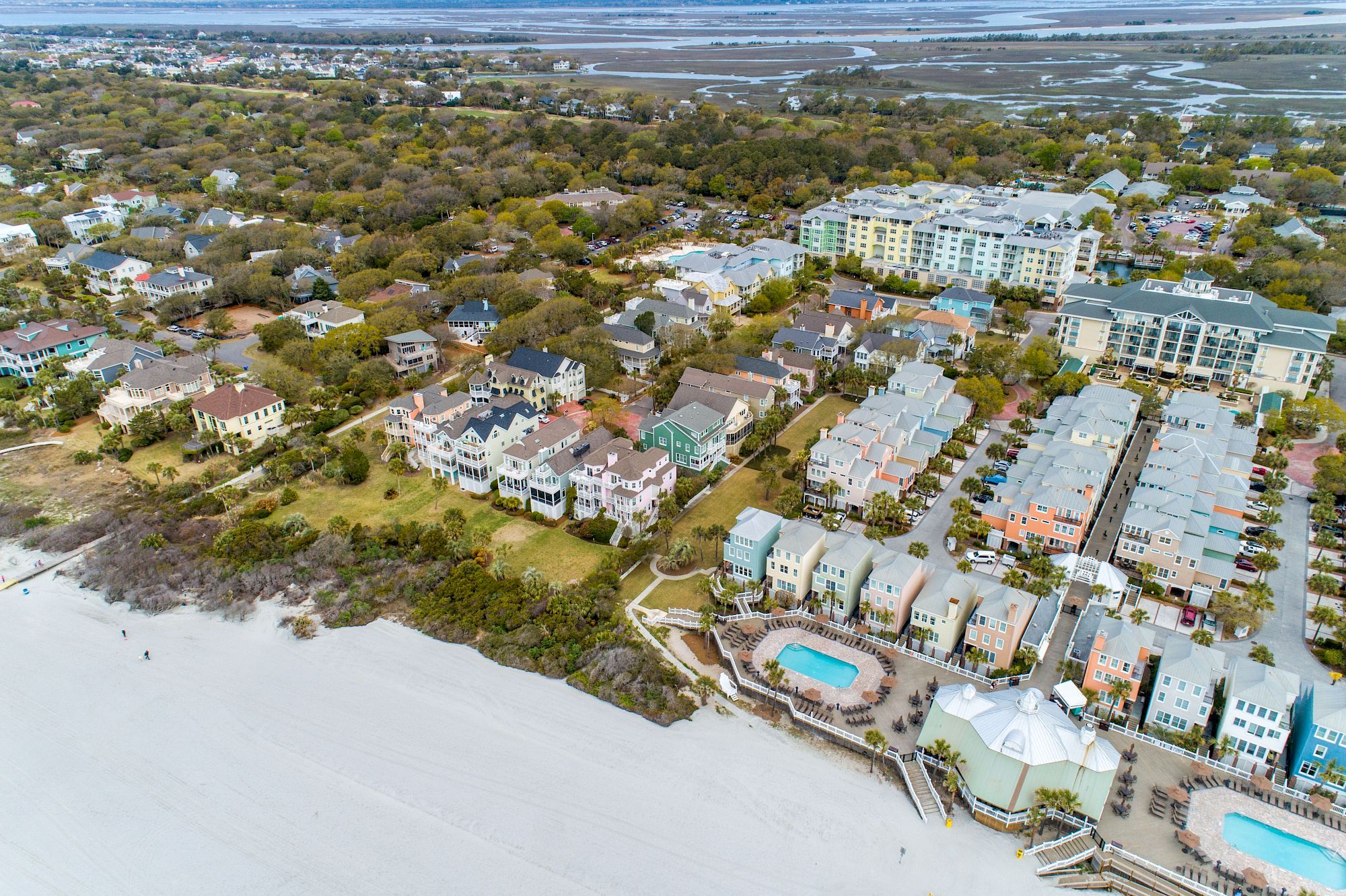 An aerial view of a coastal community features colorful houses, pools, and greenery near the beach with vast stretches of sandy shore.