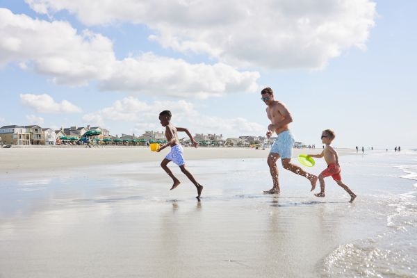A man and two kids run joyfully on a sunny beach, carrying toys, splashing through shallow water.