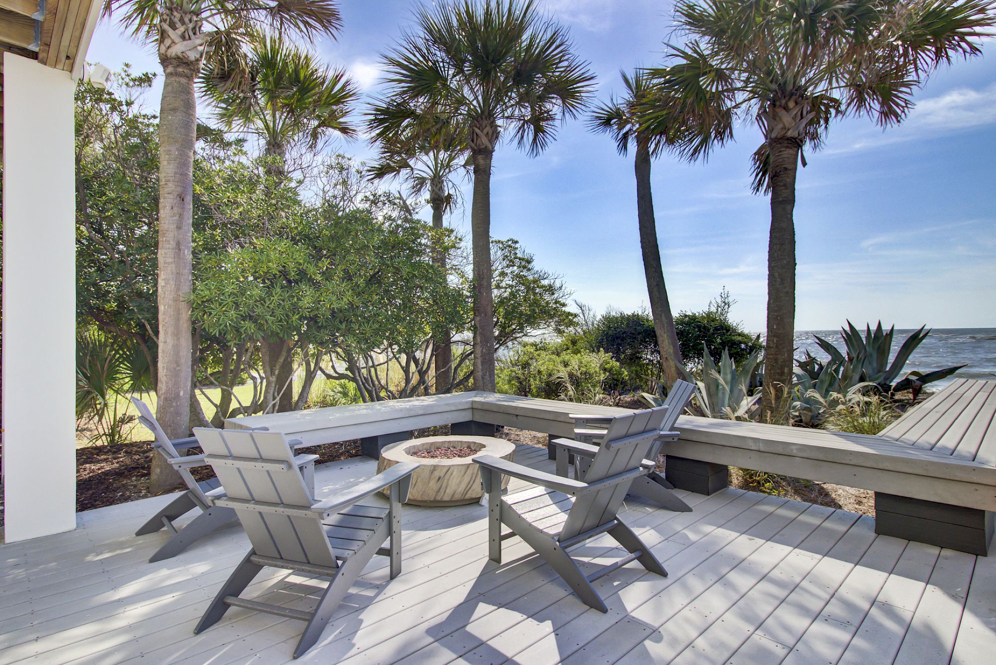 A wooden deck with chairs and a fire pit, surrounded by palm trees, overlooking the ocean under a clear blue sky.