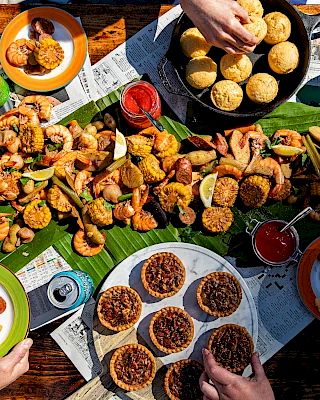 A festive table with seafood, small cakes, canned drinks, and people reaching in to eat. The setup includes banana leaves as decoration.
