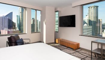 The image shows a modern hotel room with large windows, a bed, a wall-mounted TV, and a view of a city skyline with tall buildings.
