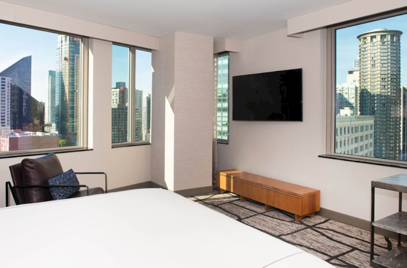 The image shows a modern hotel room with large windows, a bed, a wall-mounted TV, and a view of a city skyline with tall buildings.