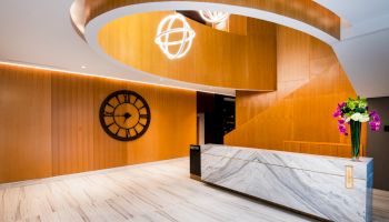 A modern lobby with a wooden spiral staircase, marble reception desk, large clock, and decorative lights and flowers.